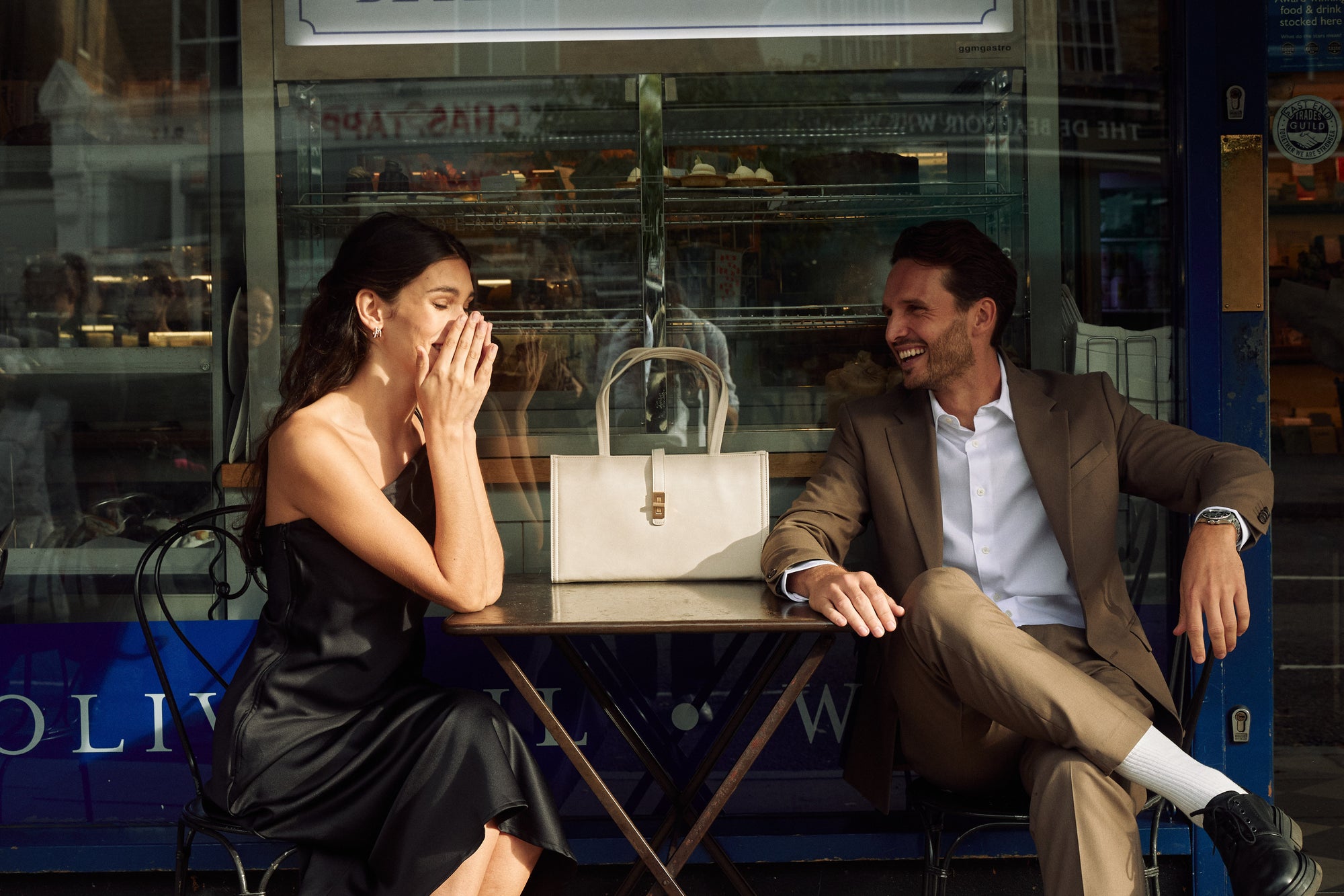 A couple laughing together outside a neighbourhood café in London with an ivory MJade handbag.