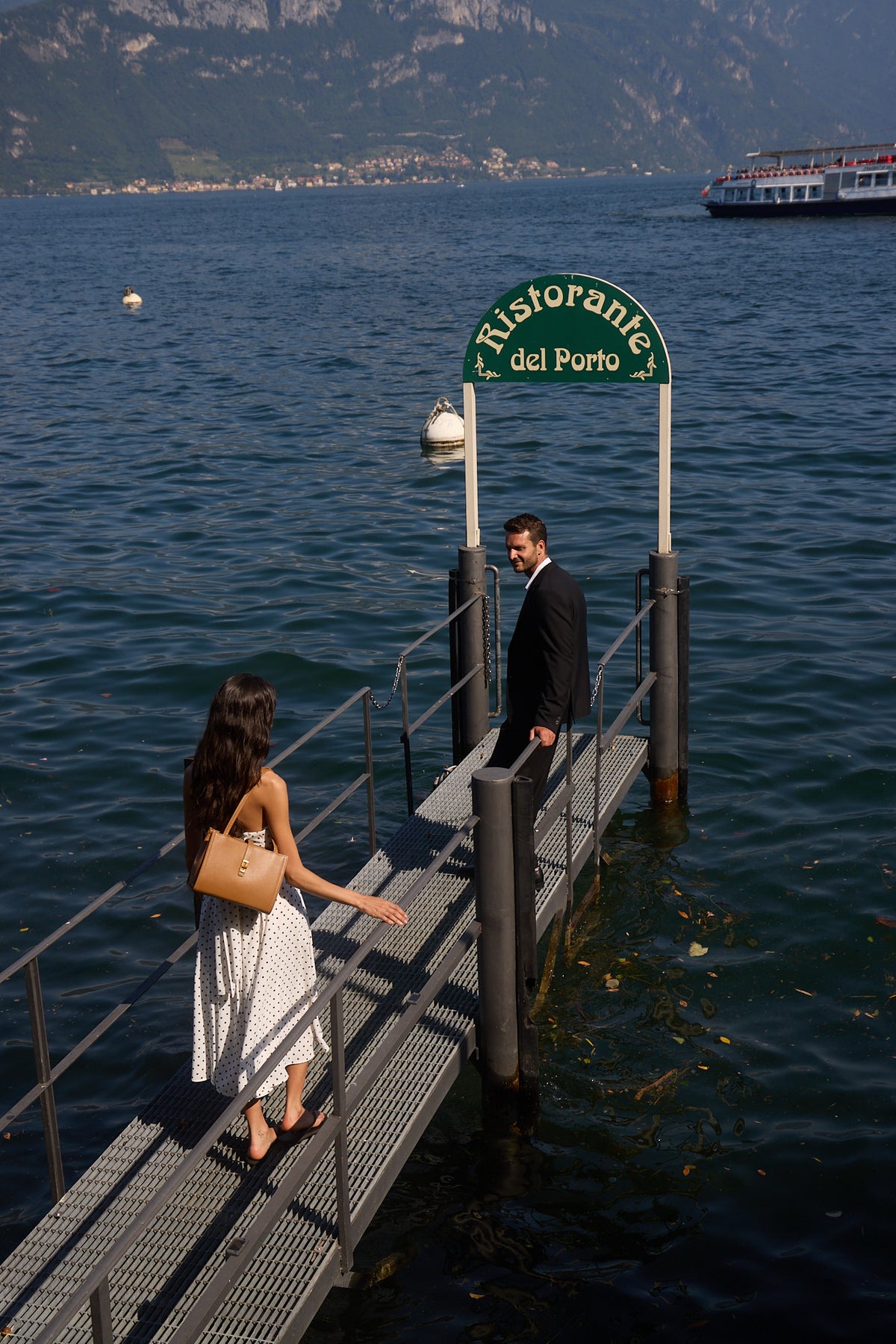 A woman carrying a tan MJade handbag approaches a man at a Lake Como landing.